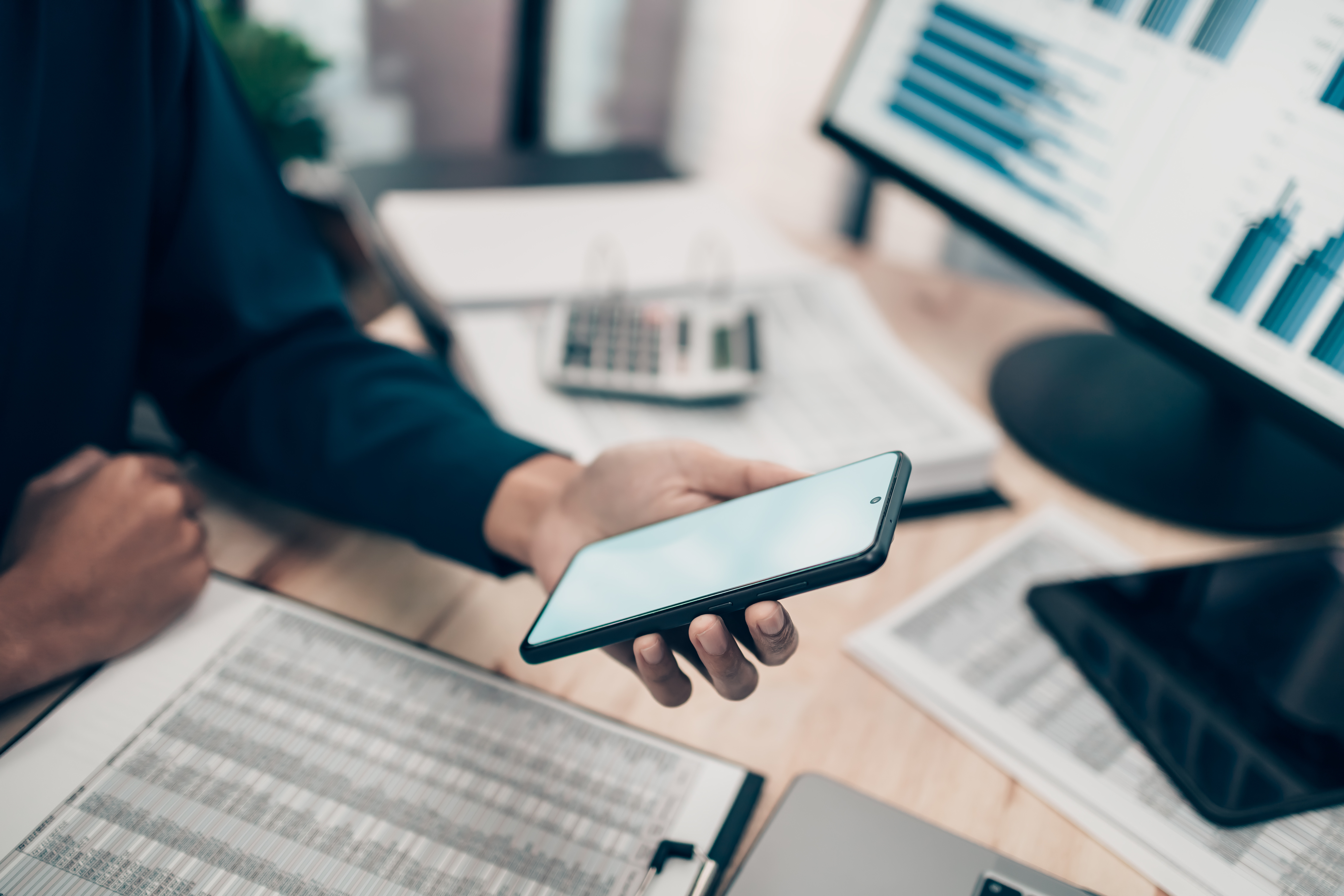Hand holding phone with charts and computer screen in background